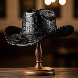 May include: A black cowboy hat with a textured, crocodile-like pattern. The hat is displayed on a wooden hat stand. The hat has a wide brim and a braided band around the crown. The hat is made of a shiny material.
