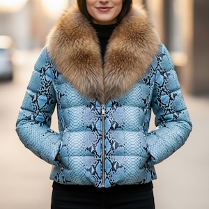 May include: A light blue puffer jacket with a snakeskin pattern and a brown fur collar. The jacket has a zipper closure and pockets. The model is wearing a black turtleneck.
