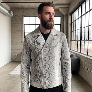 May include: A motorcycle jacket featuring a grey and white snakeskin pattern. The jacket has a notched lapel, a diagonal silver-tone metal zipper closure, and a zippered chest pocket. It is worn over a plain dark-colored shirt.