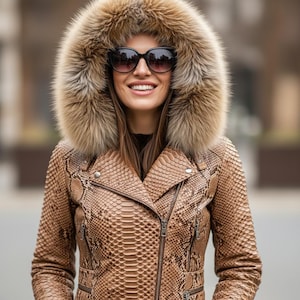 Peut inclure: Une femme porte une veste en cuir marron à motif peau de serpent avec une capuche bordée de fourrure et des lunettes de soleil. La veste a une fermeture éclair et un design de style motard. La capuche en fourrure est de couleur marron clair.