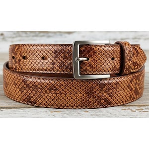 Puede incluir: Cinturón de cuero marrón con estampado de piel de serpiente. El cinturón presenta una hebilla rectangular de color plateado y una trabilla a juego. El estampado de piel de serpiente es una mezcla de tonos marrones claros y oscuros, creando una apariencia texturizada.