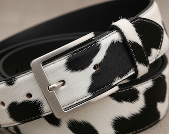 Puede incluir: Un cinturón de piel de vaca con un estampado de manchas blancas y negras. El cinturón tiene una hebilla rectangular de color plateado y un respaldo de cuero negro. El cinturón mide aproximadamente 3,8 cm de ancho.