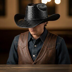 May include: A black cowboy hat with a textured, crocodile-like pattern. The hat has a wide brim and a braided band around the crown. The hat is worn by a person wearing a brown leather vest and a denim shirt.