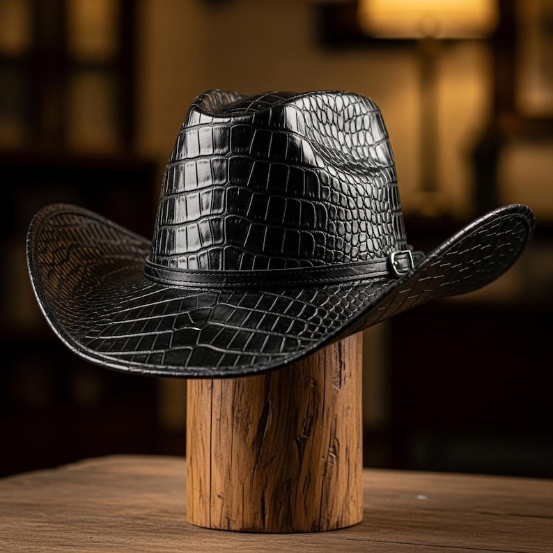 May include: A black cowboy hat with a textured, crocodile-like pattern. The hat features a wide brim and a leather band with a silver buckle. It is displayed on a wooden stand, showcasing its shape and design.
