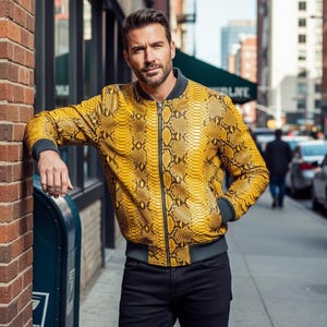 May include: A yellow and brown snakeskin-patterned bomber jacket with a black zipper and gray trim. The jacket has two pockets and is worn with black pants. The jacket is a stylish outerwear piece.