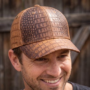 Puede incluir: Una gorra de béisbol con un patrón de textura de piel de cocodrilo en tonos bronceado y marrón. La gorra presenta una corona estructurada con paneles cosidos, una visera curva y pequeños ojales metálicos en los laterales de la pieza.