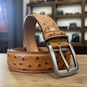 Puede incluir: Un cinturón de cuero marrón claro con una hebilla plateada. El cinturón tiene una superficie texturizada con pequeños puntos marrones oscuros. La hebilla rectangular tiene un acabado de metal cepillado. El cinturón está enrollado sobre una superficie de madera.