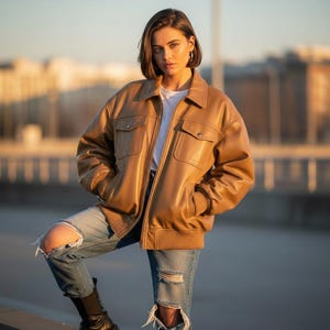 Peut inclure: Veste en cuir marron clair avec col, deux poches poitrine et poignets côtelés. La veste est portée sur un t-shirt blanc et associée à un jean bleu délavé et des bottes noires.