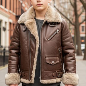 Może przedstawiać: Brązowa skórzana kurtka bomberka z beżowym kołnierzem i podszewką z owczej skóry. Kurtka posiada zapięcie na zamek błyskawiczny, wiele kieszeni i detale z klamrami na mankietach. Kurtka dla dzieci.