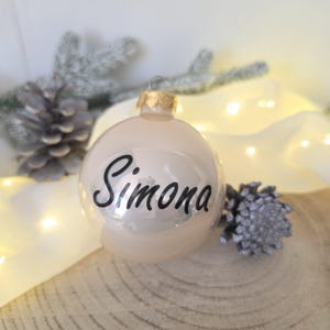 May include: A spherical, glossy, cream-colored Christmas ornament featuring the name Simona written in black cursive script. The ornament has a gold-toned metal cap at the top for hanging, providing a reflective surface that catches light.