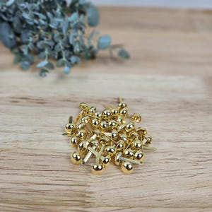 May include: A collection of small, metallic gold-colored paper fasteners, each featuring a smooth, rounded dome head and two flat, flexible prongs extending from the base, arranged in a loose pile on a light-toned wooden surface.