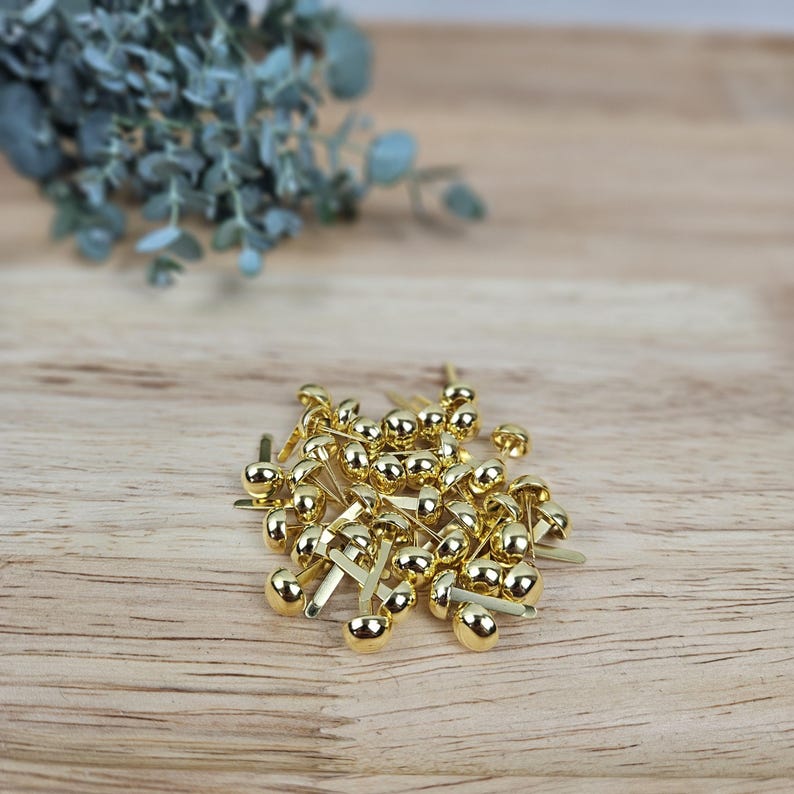 May include: A collection of small, metallic gold-colored paper fasteners, each featuring a smooth, rounded dome head and two flat, flexible prongs extending from the base, arranged in a loose pile on a light-toned wooden surface.