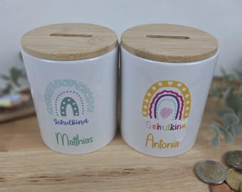 May include: Two white ceramic coin banks with light brown bamboo lids featuring a coin slot. Each bank displays a colorful rainbow graphic, the German text Schulkind, and the names Matthias and Antonia printed in green and yellow respectively.