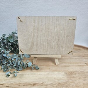 May include: A rectangular light-colored wooden board held by a small wooden easel. The board features four gold-toned metal clips positioned at each corner to secure items. The wood grain is visible across the surface of the board and the easel.