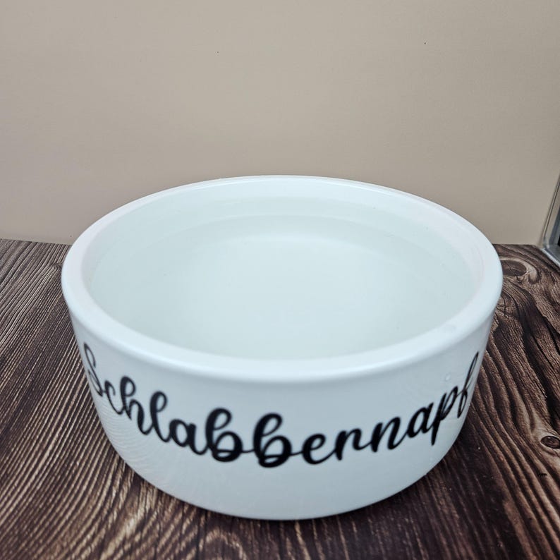 Op de afbeelding: Een witte keramische voerbak voor huisdieren gevuld met water. De kom heeft een afgeronde vorm en een brede opening. Zwarte cursieve tekst met de tekst "Schlabbernapf" staat op de zijkant van de kom. De kom staat op een houten oppervlak.