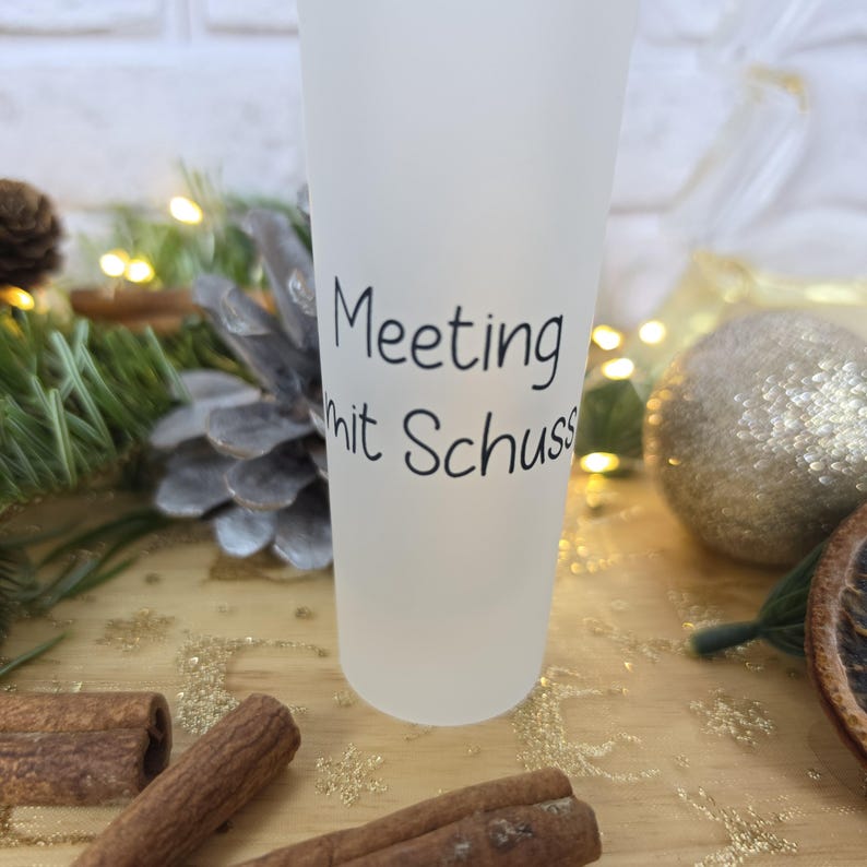 May include: A frosted glass tumbler featuring the black printed text Meeting mit Schuss. The cylindrical vessel has a smooth, semi-opaque surface finish and stands upright, displaying clear, legible lettering centered on the front of the glass.