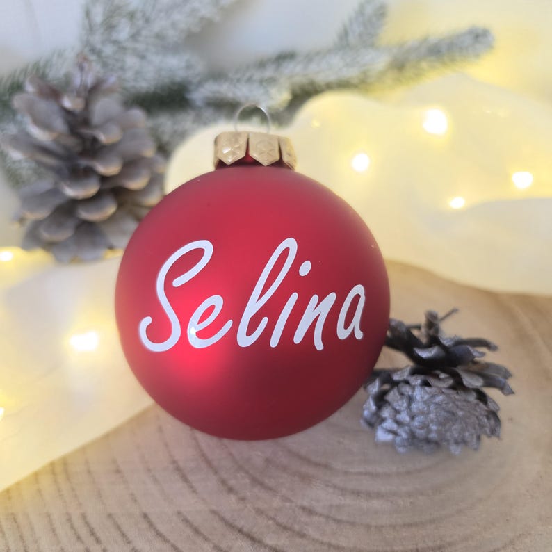 May include: A matte red spherical Christmas ornament featuring the name Selina written in white cursive script. The ornament has a metallic gold cap at the top and is positioned centrally, displaying a smooth, solid-colored surface finish.