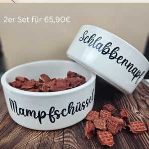 Op de afbeelding: Twee witte keramische voerbakken voor huisdieren met zwarte cursieve tekst. E&eacute;n bak is gevuld met bruin dierenvoer. De tekst op de bakken luidt "Mampfsch&uuml;ssel" en "Schlabbernapf". De bakken staan op een houten oppervlak.