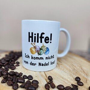 以下が含まれることがあります： ドイツ語のテキスト「Hilfe! Ich komm nicht von der Nadel los!」と毛糸玉と針のグラフィックが描かれた白いセラミックマグ。マグカップは、コーヒー豆が置かれた木製の表面に置かれています。