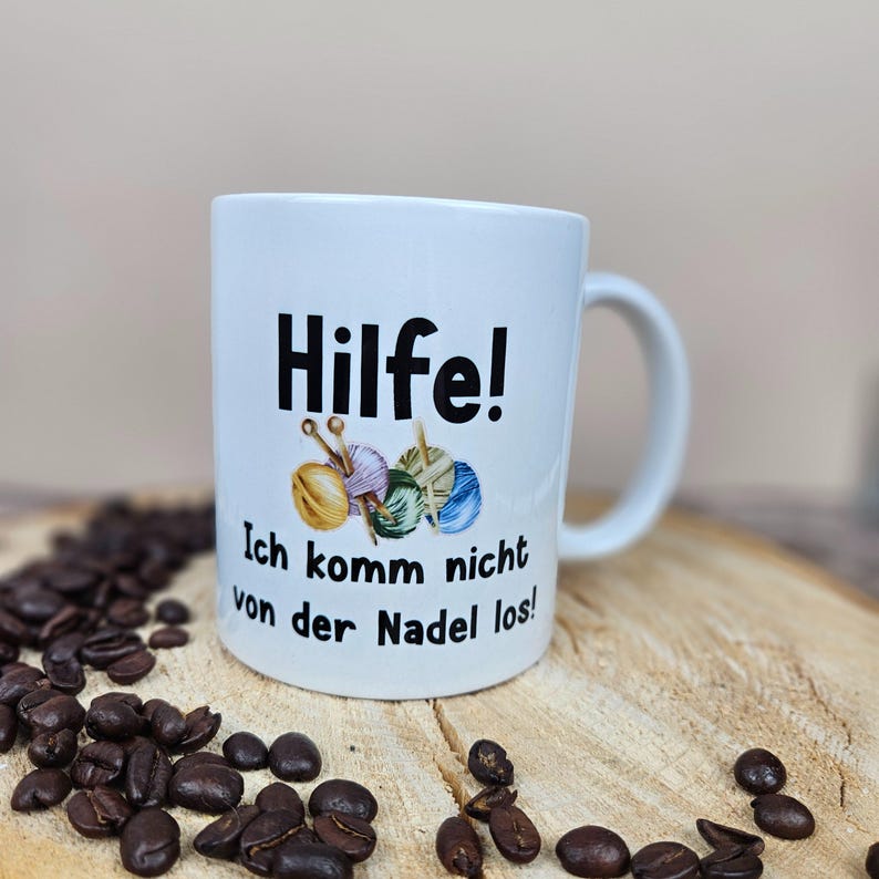 以下が含まれることがあります： ドイツ語のテキスト「Hilfe! Ich komm nicht von der Nadel los!」と毛糸玉と針のグラフィックが描かれた白いセラミックマグ。マグカップは、コーヒー豆が置かれた木製の表面に置かれています。