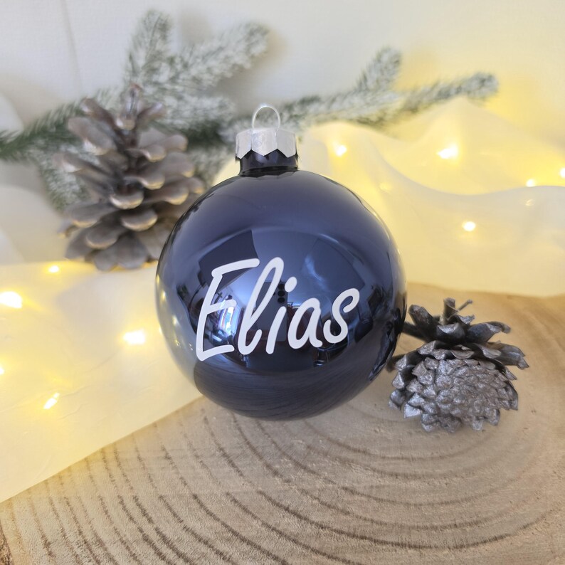 May include: A glossy dark navy blue spherical Christmas ornament featuring the name Elias written in white cursive script. The ornament has a silver-toned metal cap at the top for hanging and a smooth, reflective surface finish.