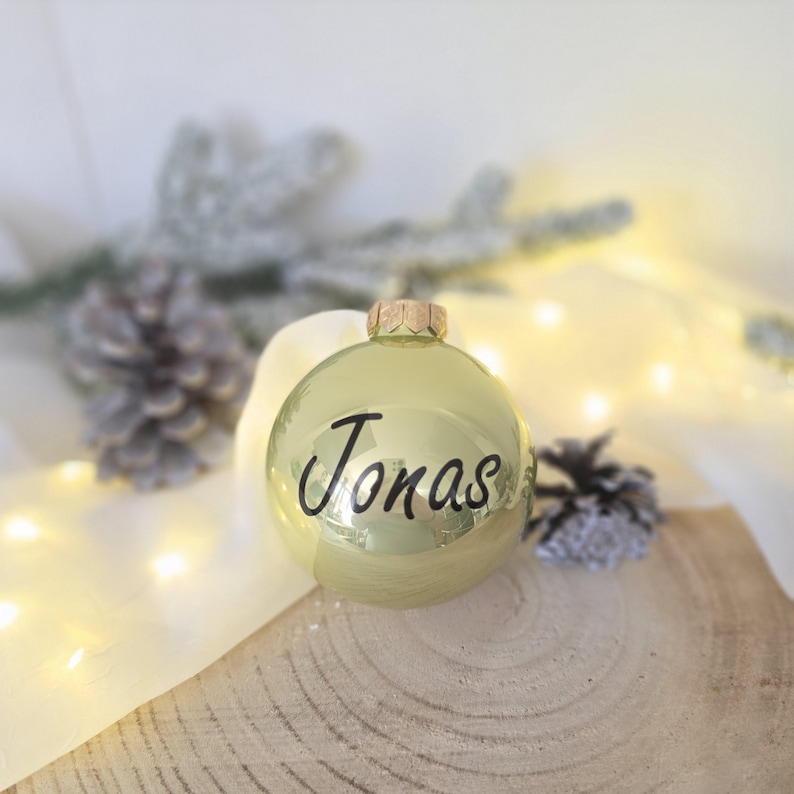May include: A spherical, shiny gold Christmas ornament featuring the name Jonas written in black cursive script. The ornament has a metallic gold cap at the top and a smooth, reflective surface that catches the surrounding light.