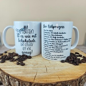 May include: Two white ceramic mugs with black text and coffee beans on a wooden surface. One mug reads "Mit Stoffen ist es wie mit Schokolade, EIN STUCK IST NIE genug!" The other mug lists steps in German.