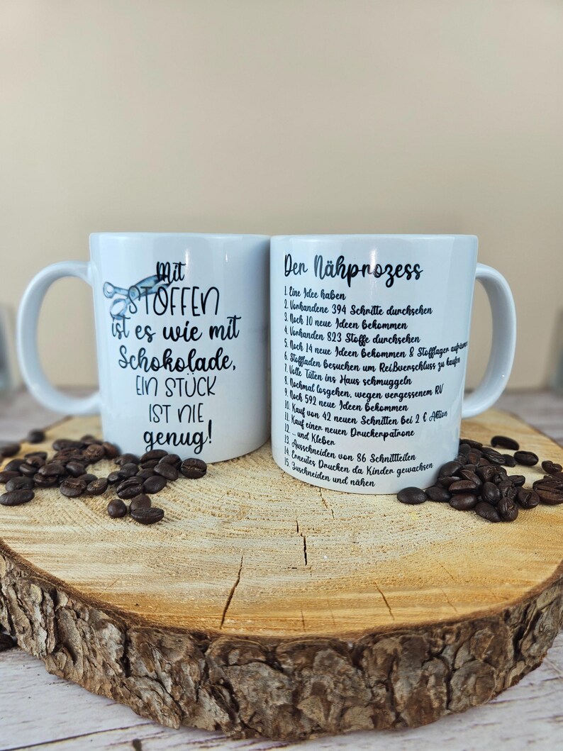 May include: Two white ceramic mugs with black text and coffee beans on a wooden surface. One mug reads "Mit Stoffen ist es wie mit Schokolade, EIN STUCK IST NIE genug!" The other mug lists steps in German.