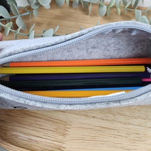 May include: A light gray felt pencil case with a zipper, open to reveal several colored pencils. The pencils are orange, yellow, purple, green, and blue. The case is on a wooden surface.