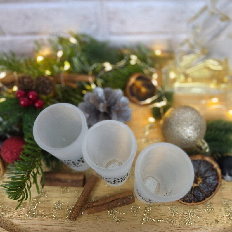 May include: Three frosted glass shot glasses with visible black text on the exterior. The glasses are cylindrical with a smooth rim and a clear base, arranged in a cluster on a wooden surface surrounded by pine branches, cinnamon sticks, and ornaments.