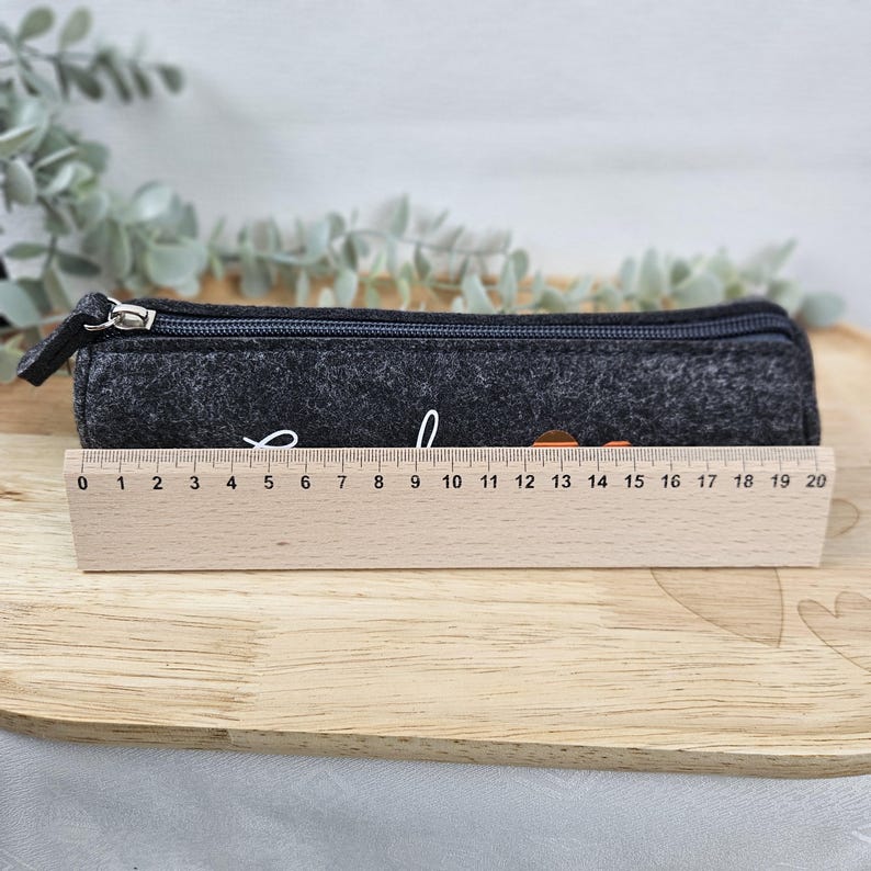 May include: A dark gray felt pencil case with a zipper closure, measuring approximately 8 inches in length. The pencil case has white cursive text and a small orange design. It rests on a wooden tray with a wooden ruler.