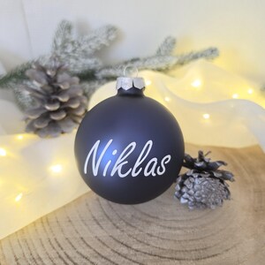 May include: A matte black spherical Christmas ornament featuring the name Niklas written in white cursive script. The ornament has a silver-toned metal cap at the top for hanging and a smooth, solid-colored surface finish.