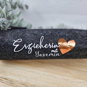 May include: A dark gray felt pencil case with the German word "Erzieherin" in white script, the name "Yasemin" in white, and a copper-colored heart. The pencil case is on a wooden surface.