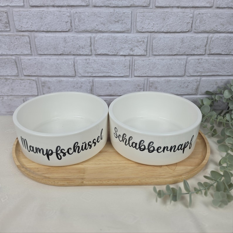 Pode incluir: Dois recipientes de cer&acirc;mica branca para animais de estima&ccedil;&atilde;o com texto em cursivo preto. O recipiente da esquerda tem a etiqueta Mampfsch&uuml;ssel e o da direita Schlabbernapf. Os recipientes est&atilde;o lado a lado numa bandeja de madeira oval.