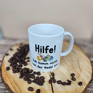以下が含まれることがあります： ドイツ語のテキスト「Hilfe! Ich komm nicht von der Nadel los!」と糸と針のグラフィックが描かれた白いセラミックマグ。マグカップは木製の表面にコーヒー豆で囲まれています。