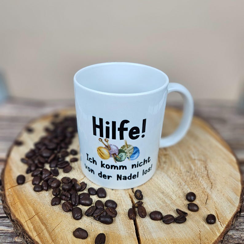 以下が含まれることがあります： ドイツ語のテキスト「Hilfe! Ich komm nicht von der Nadel los!」と糸と針のグラフィックが描かれた白いセラミックマグ。マグカップは木製の表面にコーヒー豆で囲まれています。