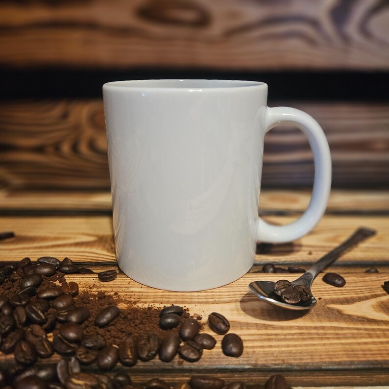 May include: A white ceramic coffee mug with a curved handle sits on a wooden surface. Coffee beans and ground coffee surround the mug, with a spoon holding a few beans. The background is a wooden panel.