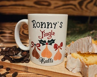 May include: White ceramic mug with the text "Ronny's Jingle Balls" in black and red script. The mug features illustrations of three ornaments with bows. Coffee beans and sliced cake are also visible.