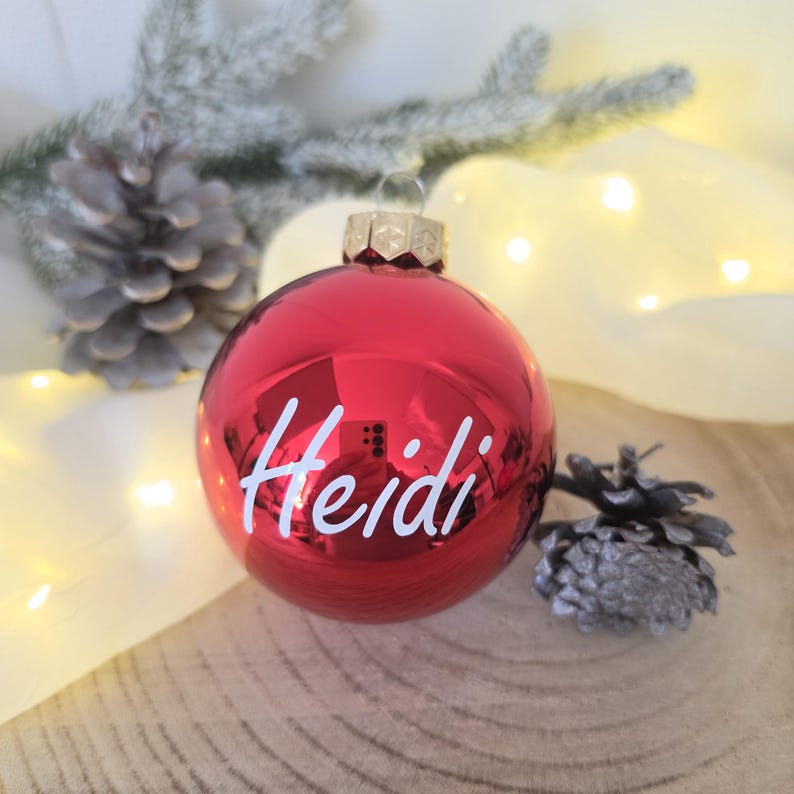 May include: A shiny red spherical Christmas ornament featuring the name Heidi written in white cursive script. The ornament has a gold-colored metal cap at the top and a reflective surface that shows a faint reflection of a smartphone camera.
