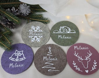 May include: Five circular felt coasters in gray, green, purple, brown, and mauve. Each coaster features a white graphic and the name Melanie. Designs include a snowflake, a cat, bells, a Christmas tree, and a deer head with a small heart.