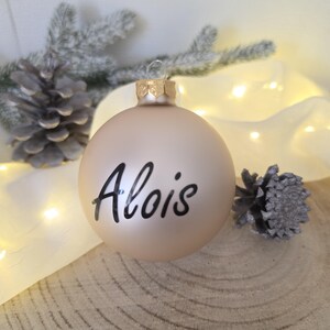 May include: A matte champagne-colored spherical Christmas ornament featuring the name Alois written in black script lettering. The ornament has a metallic gold-toned cap at the top with a small loop for hanging, centered in the composition.