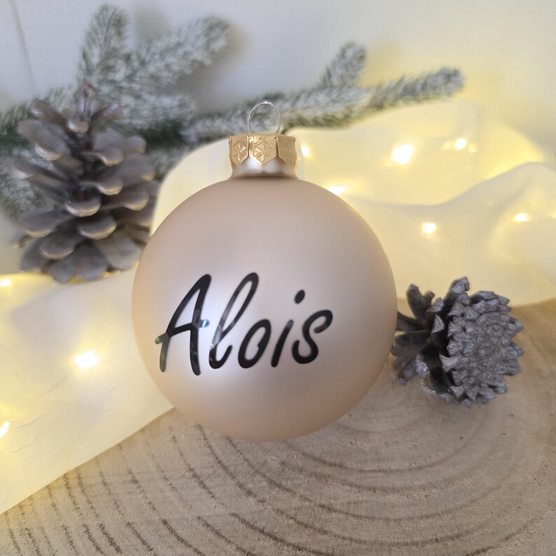 May include: A matte champagne-colored spherical Christmas ornament featuring the name Alois written in black script lettering. The ornament has a metallic gold-toned cap at the top with a small loop for hanging, centered in the composition.