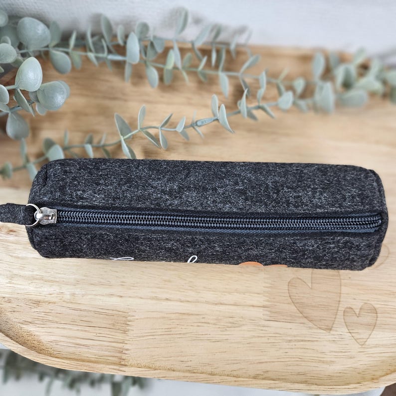 May include: A dark gray felt pencil case with a zipper closure. The rectangular case has a small loop on one end and a silver zipper. The case is on a wooden tray with decorative leaves in the background.