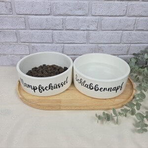 Pode incluir: Dois recipientes de cer&acirc;mica branca para animais de estima&ccedil;&atilde;o sobre uma bandeja oval de madeira. O recipiente esquerdo cont&eacute;m ra&ccedil;&atilde;o seca castanho-escura e tem o texto preto Mampfsch&uuml;ssel. O direito est&aacute; cheio de &aacute;gua e tem o texto preto Schlabbennapf.