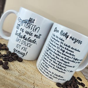 May include: Two white ceramic mugs with black text and illustrations. One mug reads "Mit STOFFEN ist es wie mit Schokolade, EIN ST&Uuml;CK IST NIE genug!" The other mug lists steps in German. Coffee beans are scattered around the mugs.