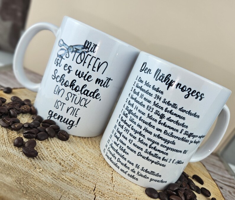 May include: Two white ceramic mugs with black text and illustrations. One mug reads "Mit STOFFEN ist es wie mit Schokolade, EIN ST&Uuml;CK IST NIE genug!" The other mug lists steps in German. Coffee beans are scattered around the mugs.