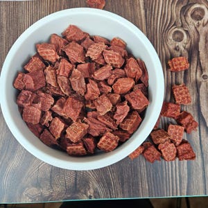 Op de afbeelding: Een witte kom gevuld met roodbruine, in blokjes gesneden hondenkoekjes. Enkele traktaties liggen verspreid op een houten oppervlak. De traktaties hebben een gestructureerd, rasterachtig patroon aan sommige kanten.