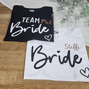 K&ouml;nnte beinhalten: Ein schwarzes T-Shirt mit wei&szlig;em Text TEAM Mel und Bride mit einem Herz sowie ein wei&szlig;es T-Shirt mit schwarzem Text Steffi und Bride mit einem Herz. Zwei klare Glasfl&ouml;ten befinden sich in der N&auml;he der T-Shirts auf einer Holzoberfl&auml;che.