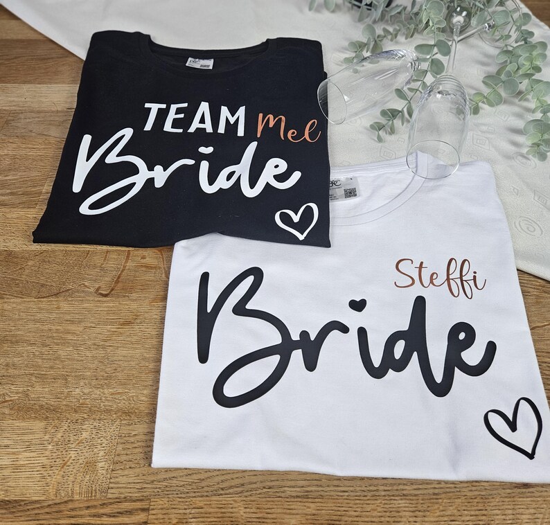 K&ouml;nnte beinhalten: Ein schwarzes T-Shirt mit wei&szlig;em Text TEAM Mel und Bride mit einem Herz sowie ein wei&szlig;es T-Shirt mit schwarzem Text Steffi und Bride mit einem Herz. Zwei klare Glasfl&ouml;ten befinden sich in der N&auml;he der T-Shirts auf einer Holzoberfl&auml;che.