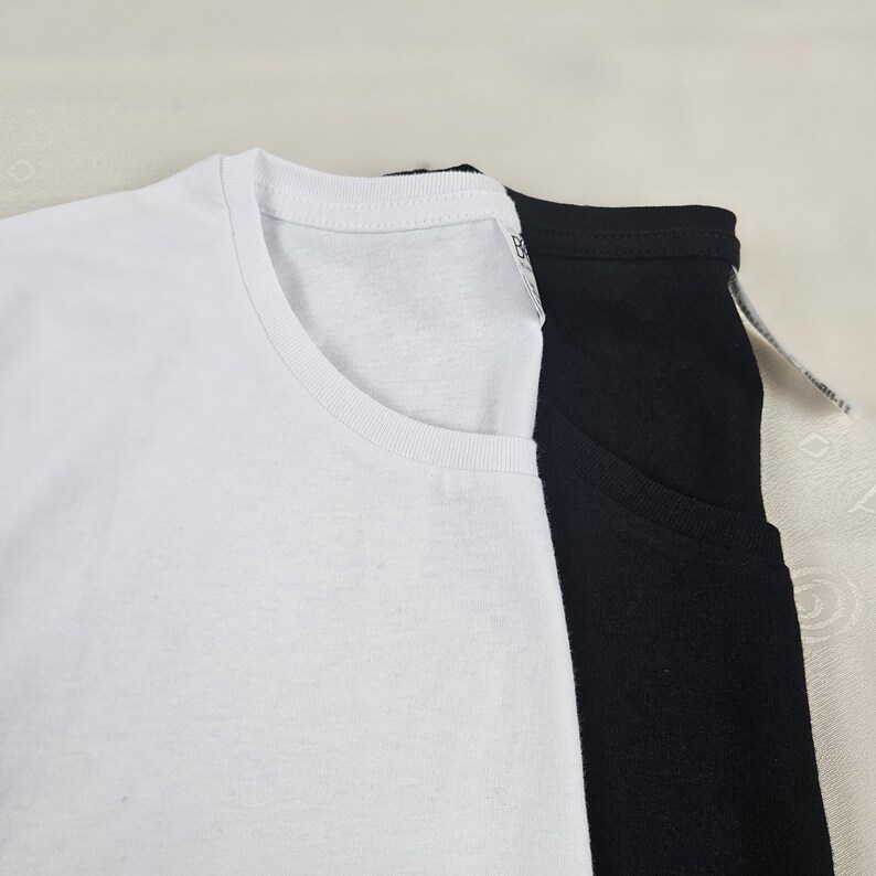 K&ouml;nnte beinhalten: Ein wei&szlig;es T-Shirt mit Rundhalsausschnitt, das &uuml;ber einem schwarzen T-Shirt mit Rundhalsausschnitt liegt. Beide T-Shirts haben einen gerippten Kragen und ein kleines wei&szlig;es Stoffetikett an der Innenseite des Ausschnitts. Das wei&szlig;e T-Shirt liegt im Vordergrund und &uuml;berlappt teilweise das schwarze T-Shirt, wobei der einfarbige Stoff beider Kleidungsst&uuml;cke sichtbar ist.
